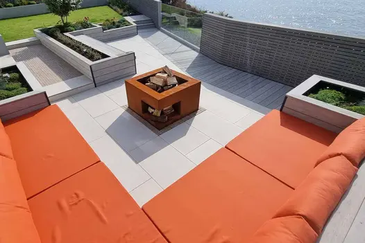 Sea views seen from multi level garden using luna designboard composite decking around built in bench with orange cushions around steel corten log burner.