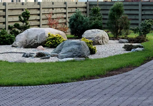 Feature of large rocks arranged on small light pebbles sits on curved lawn with small trees, flanked by path of Amersham Clay Pavers.