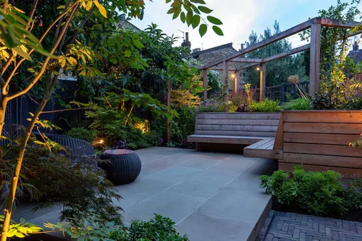 Raised patio of 900x600 Heath sawn sandstone slabs with cantilevered wooden benches and multi-stem shrubs. Design by Daniel Shea
