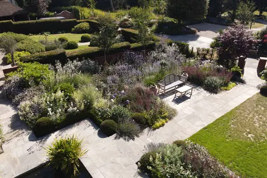 Tumbled Black Sandstone paving runs between flowerbeds and lawn, with a bench and table to the left, surrounded by trees and hedges.