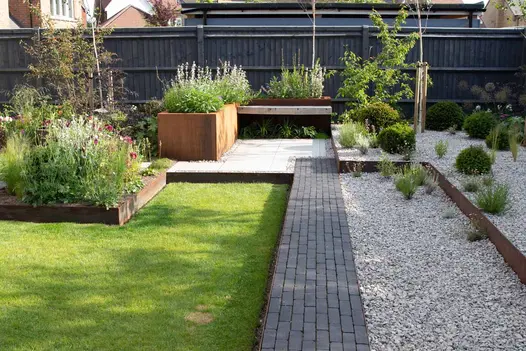 Amersham clay paver path leads to Corten steel planters brimming with green and purple plants, with light pebbles on the right side.