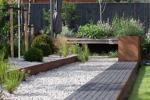 Corten steel planters filled with green and purple plants, a path of amersham clay pavers leads to it with light pebbles on the left.