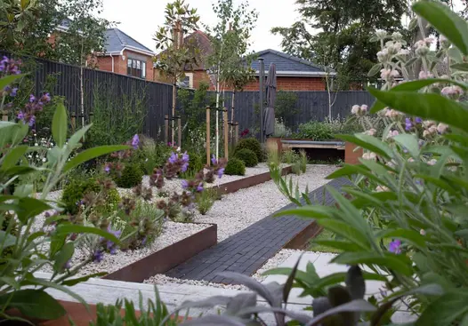 View through green and purple plants, with an Amersham clay paver path leading up to planters and light pebbles on the left.