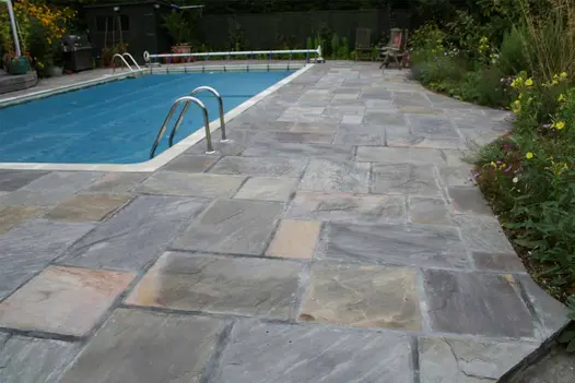 Large rectangular swimming pool with wide Tumbled Black sandstone paving surround, edged with planted border and fence.