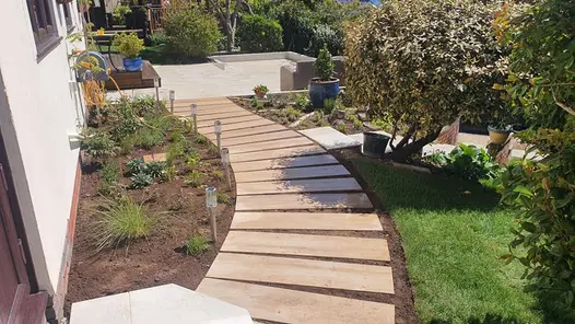 Rovere wood effect stepping stone path curving through a garden towards a patio seating area with plants and grass either side of the path.