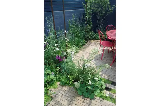 Red metal furniture sits in corner of garden on Westminster clay paving edged with planted border at base of tall black fence. 