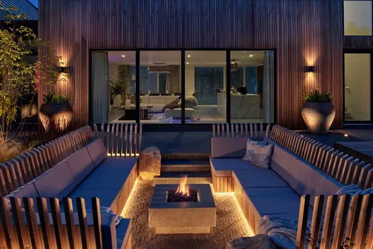 2 long inbuilt sofas either side of fire pit on Amersham clay paving in front of illuminated room with bifold doors.