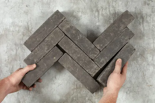 Two hands hold amersham clay paving in view from above organised in herringbone pattern with nine brick pavers on blank background.