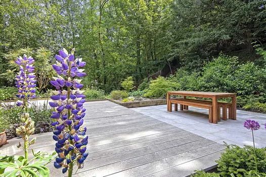 Smoked Oak Millboard Decking in woodland, with planted beds. Step down to table and benches on paving. Design by Andrea Newill.