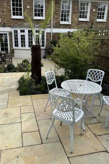 White metal table and four chairs sit on Antique Yellow Limestone Paving, withoutset planting areas, house in the background.