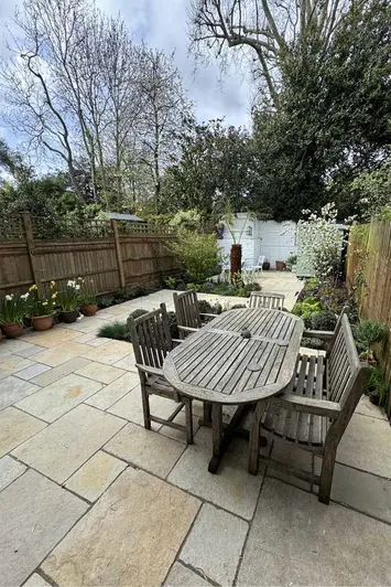 Five seat wooden table and chair set with antique yellow limestone paving in long garden with painted white brick walls.