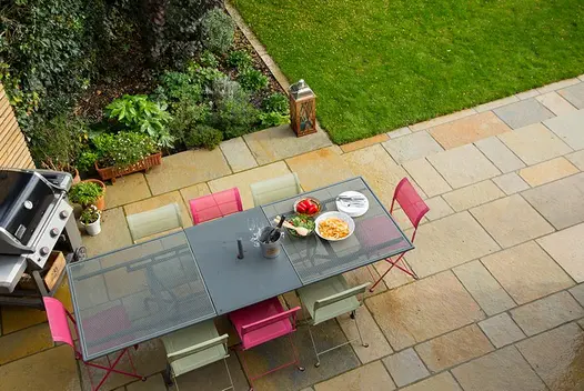 View down onto large dining set and gas barbecue on patio of Antique Yellow limestone project paving laid in courses, next to lawn.