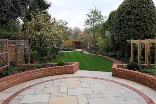 2 curve-edged beds form corners of Antique Yellow limestone patio with curved brick paver detailing in fenced garden with lawn.