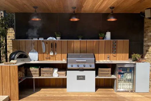 Covered outdoor kitchen with chiller and gas barbecue against wall partially clad in wood and Steel Dark external cladding.