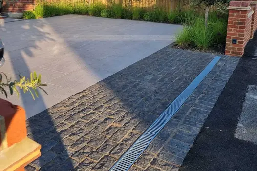 Channel drain sits between brick gate posts in wide strip of natural Black granite setts at edge of paved front garden.
