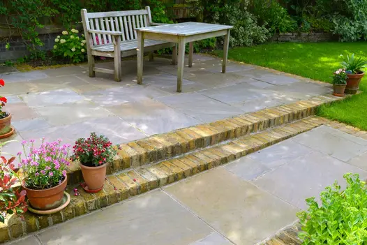 Next to garden border and fence, wooden bench and table sit on oblong patio of Autumn Brown Indian sandstone edged with clay pavers. 
