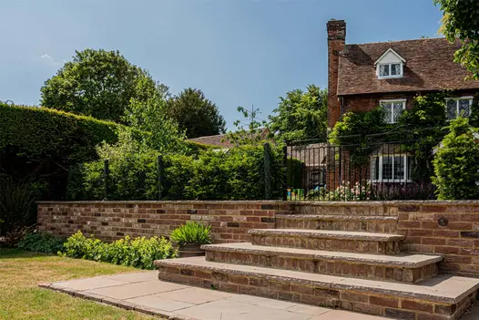5 steps up from Autumn Brown sandstone paving laid in lawn to double iron gates in low hedge with rural cottage beyond. 