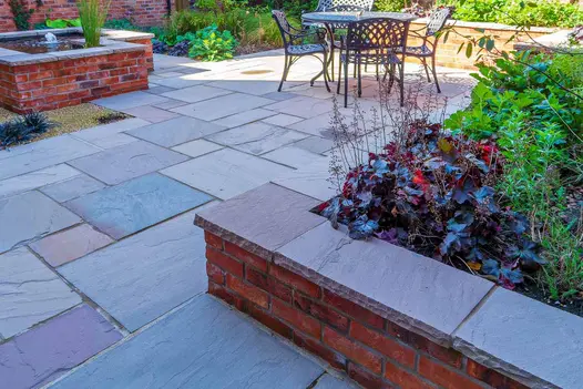Metal dining set sits on patio of Autumn Brown Indian sandstone with raised beds of brick topped with matching coping. 