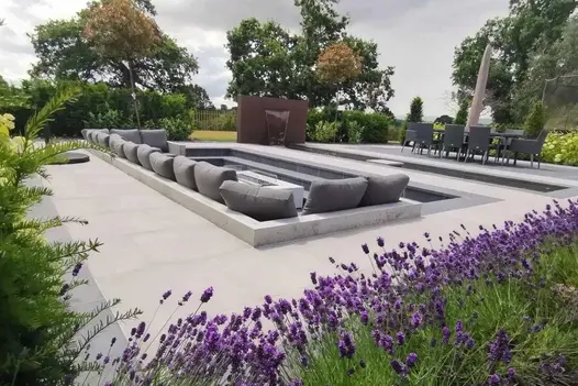 Light grey patio with sunken seating area with firepit, paved in Slab Coke porcelain paving. Design by Bancroft Lee Landscapes.