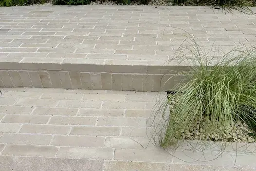 Two level patio using beige sandstone patio bricks as pavers and for cladding the step up, grassy foliage on the right.