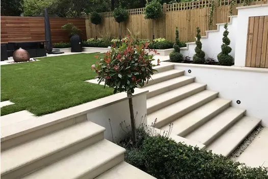 Beige Smooth Sandstone bullnose steps rise to patio with illuminated garden room and firepit past flanking upright timber sleepers.
