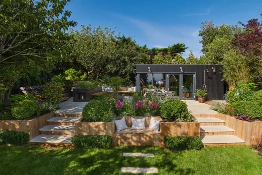 2 flights of beige smooth sandstone garden steps rise between raised beds made of sleepers to patio surrounded by shrubs and trees.