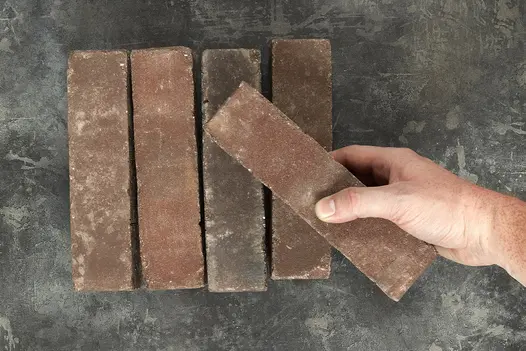 A hand places a paver at angle on top of 4 Bergamo Clay Paver Bricks showing texture and shade variations. Available nationwide.