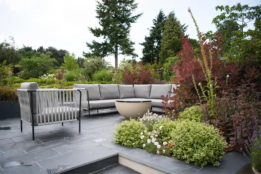 Flank wall of steps, topped with Brazilian Black Slate Copings, merges into patio with outdoor furniture. Mature garden beyond.