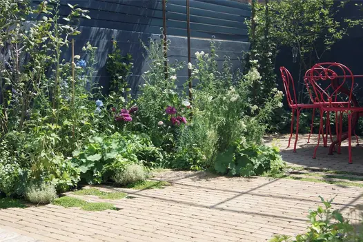 Relaxed planting in irregular-shaped bed between high dark fence and Westminster clay paving with metal furniture.