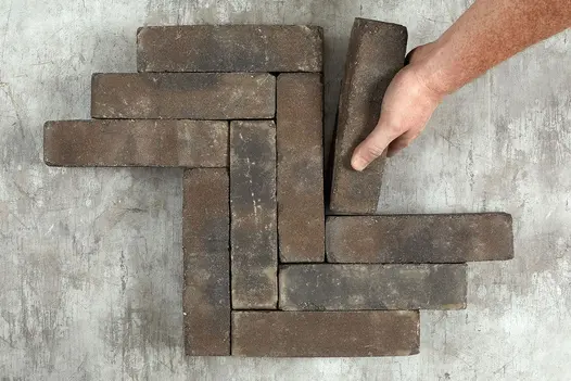 Hand places one grey and brown Ancona clay paver against 9 matching patio bricks laid in 3 zigzag rows on dark background.