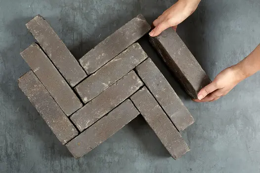 2 hands add 1 brick to 9 brown Lugano Clay Pavers in herringbone pattern on grey background. Free UK delivery available.
