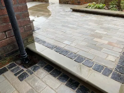 Natural Black Granite Setts laid between buff coloured stones, wet from rain, bullnose step leads up to patio.