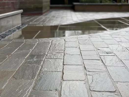 Low view of wet Kandla grey cobble setts with small puddle, showing riven surface. Design by Brockstone Landscape Construction.