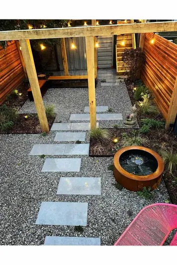 Steel Corten water feature sits next to stepping stones made from steel grey porcelain paving, running under pergola with string lights at night.