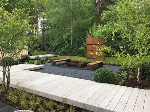 In woodland setting, raised boardwalk in Smoked Oak Millboard decking runs next to seating area with wood-topped benches.