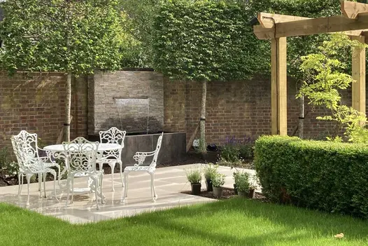 Stone-clad wall with spout pours water into trough faced with Steel Dark DesignClad next to patio with white iron table and chairs.