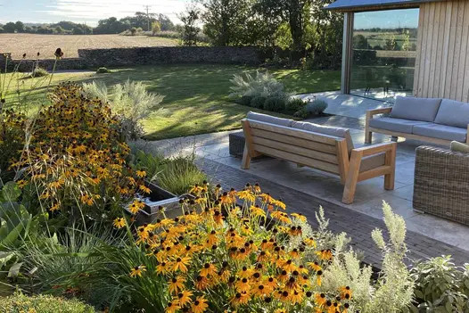 Wide strip of clay pavers frames Jura Beige sawn limestone patio between herbaceous border and wood-clad building.