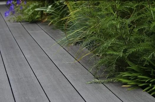 Grasses, long green leaves and blue flowers spill over edge of Charcoal composite decking boards, laid parallel to planted border.