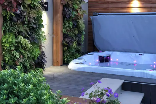 2 bullnose steps rise to open hot tub sunk into Charcoal composite decking. With slatted fencing, downlighters, and living walls.