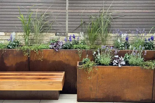 A centred profile of a tall corten steel trough planter featuring green and purple foliage, set against chestnut-toned composite battens.