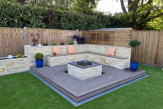 Stone-clad raised bed and bench on 2 sides of deck of Chestnut Grooved decking composite. Firepit in centre with matching cladding.