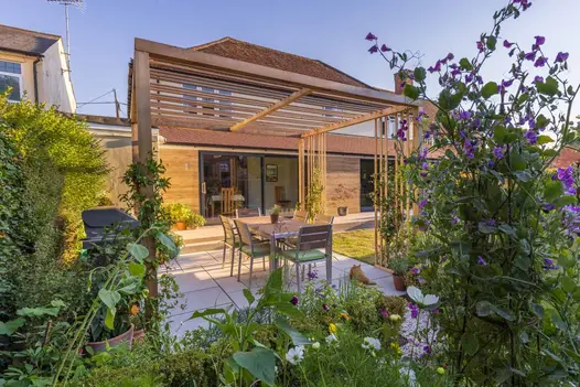 View through plants across oblong area of Dove Grey sawn sandstone paving under pergola to wide patio at back of house.