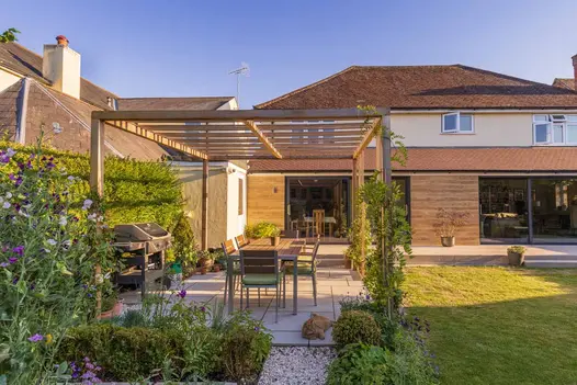 Lawn, planted bed and hedge surround patio of Dove Grey sawn sandstone paving on 3 sides, at back of house with bifold doors.