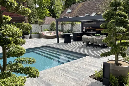 Outdoor cooking area under large pergola, with comfy seating area. Steps opposite lead down to pool, with Cinder porcelain Paving.
