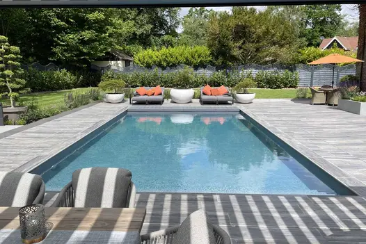 Impressive view from under pergola onlooking two sunloungers with plants either side, large pool in the middle of Cinder Porcelain Paving.