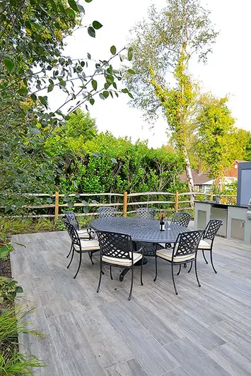Cinder porcelain patio with table and chairs and an outdoor kitchen set amongst a heavily planted garden area.