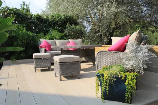 Rattan furniture with beige and pink pillows sit on Limed Oak millboard decking surrounded by trees and bushes.