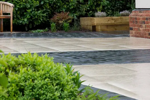 Paving of cream porcelain slabs and Midnight Black limestone cobbles, with wooden raised bed and bay laurel hedge in background.