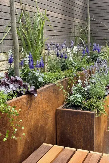 Side view of corten steel tall trough planter with green and purple foliage against chestnut coloured composite battens.