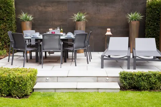 Raised patio of pale paving with Steel Dark porcelain clad wall behind. 3 tall copper-colour pots stand behind garden furniture.